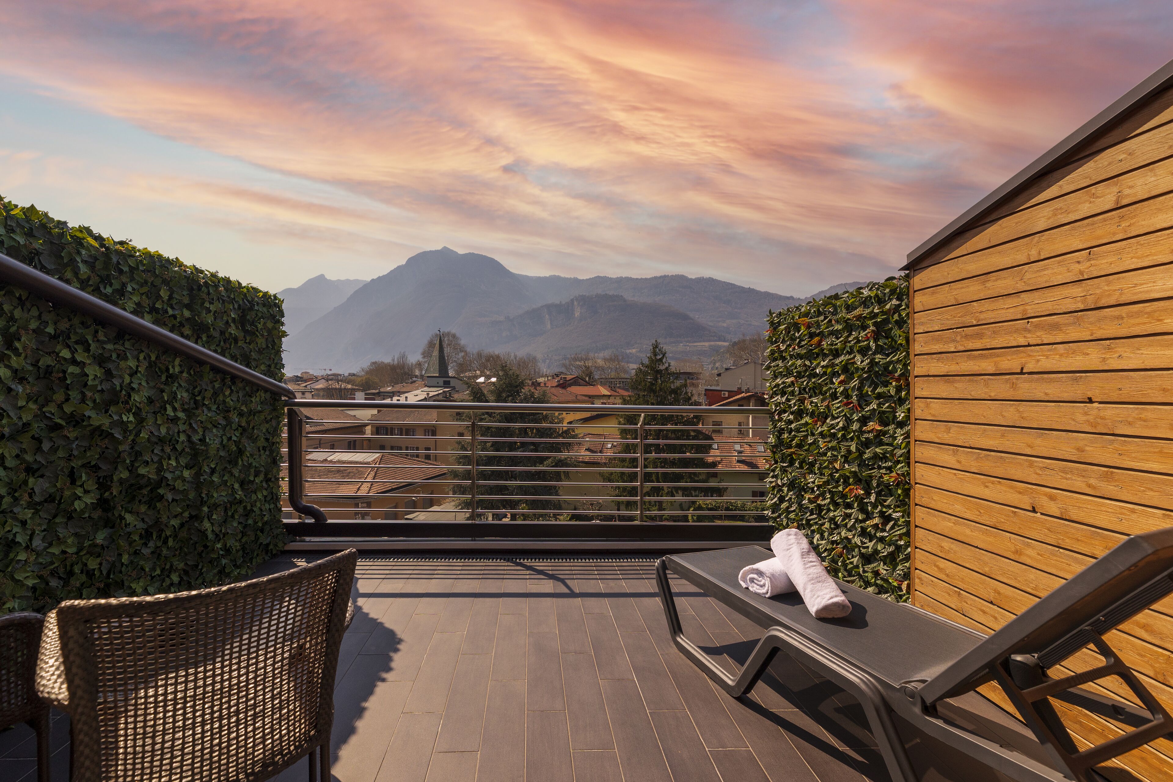A balcony with wicker chairs, a lounge chair with a towel, and a view of mountains and buildings under a colorful sky.