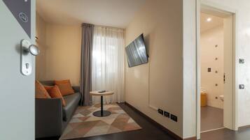 Une chambre d’hôtel moderne avec un canapé gris, une petite table ronde, un téléviseur fixé au mur et une salle de bain visible à travers une porte ouverte.