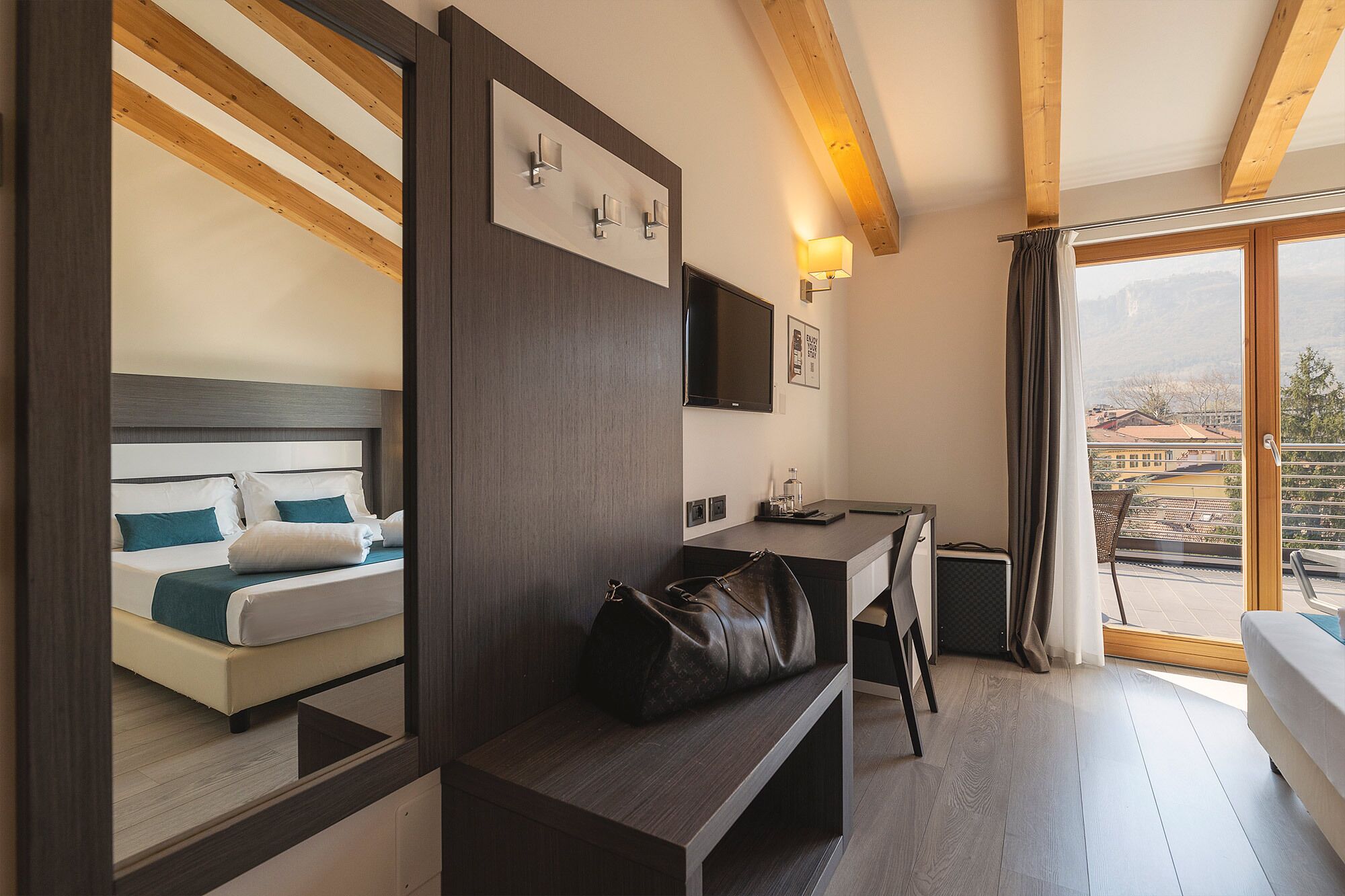 A hotel room with a bed, a desk, a bench, a TV, and a view of the city.