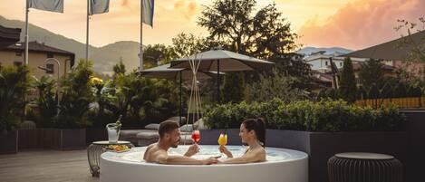 Una pareja en un jacuzzi con vista al atardecer, rodeada de mobiliario exterior y banderas.