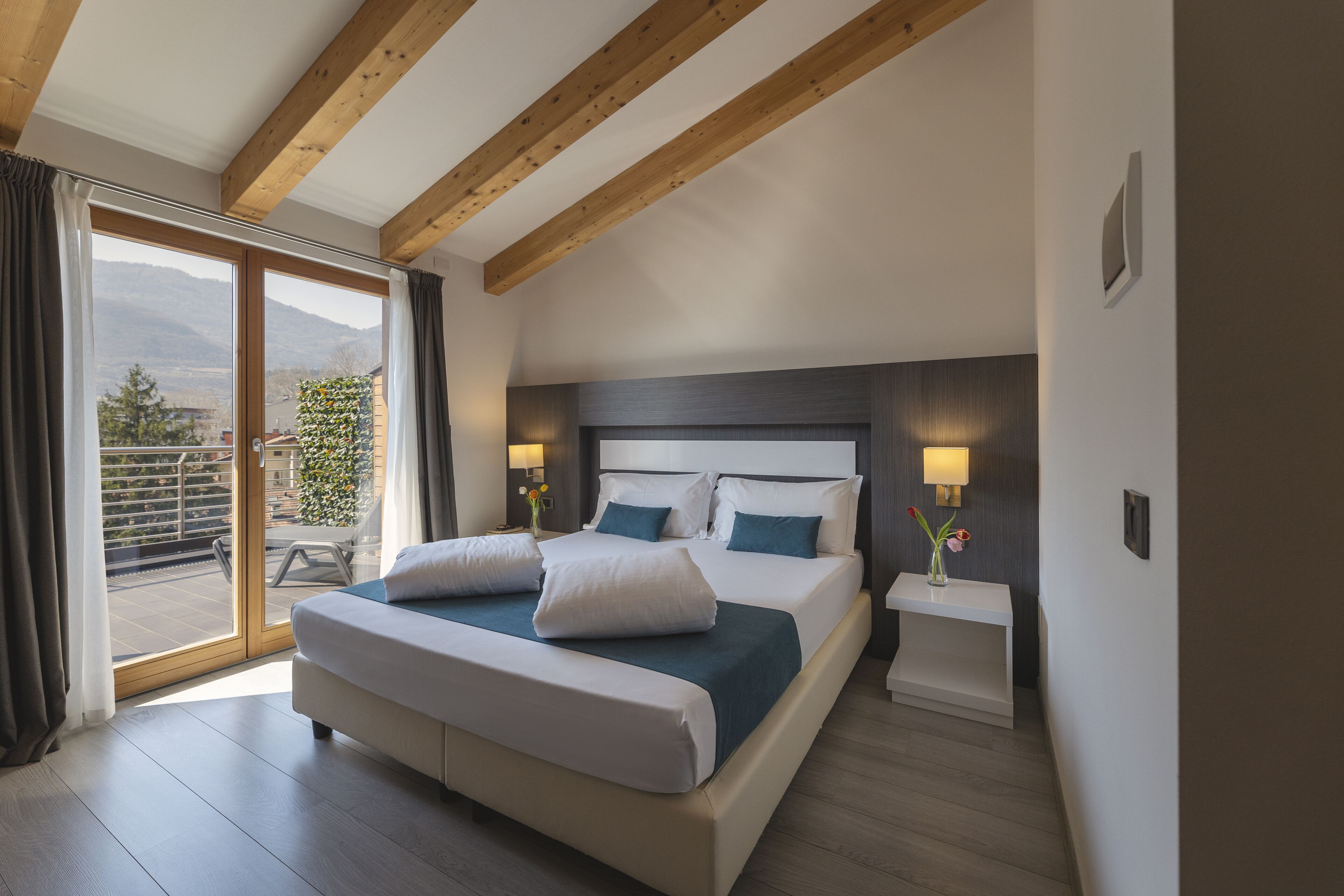Ein modernes Schlafzimmer mit einem großen Bett, Holzbalken, einem Balkon mit Blick und einem Nachttisch mit Lampe.