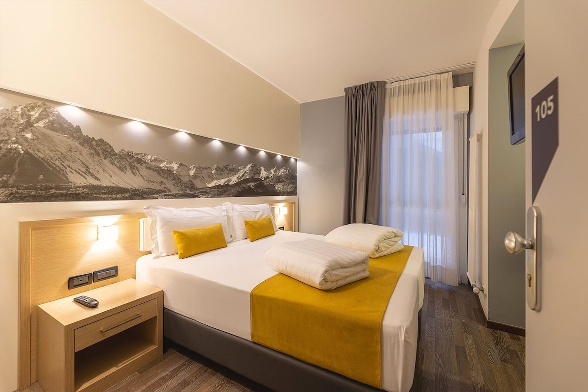 A hotel room with a bed, a nightstand, a remote control, and a mountain landscape painting on the wall.