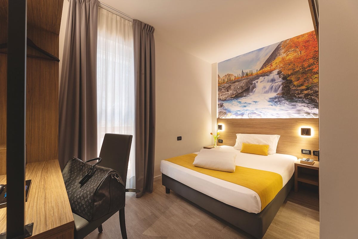 A hotel room with a bed, a chair, a desk, and a painting of a waterfall.