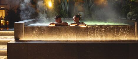 A hot tub with two people sitting inside, surrounded by tropical plants and a misty atmosphere.