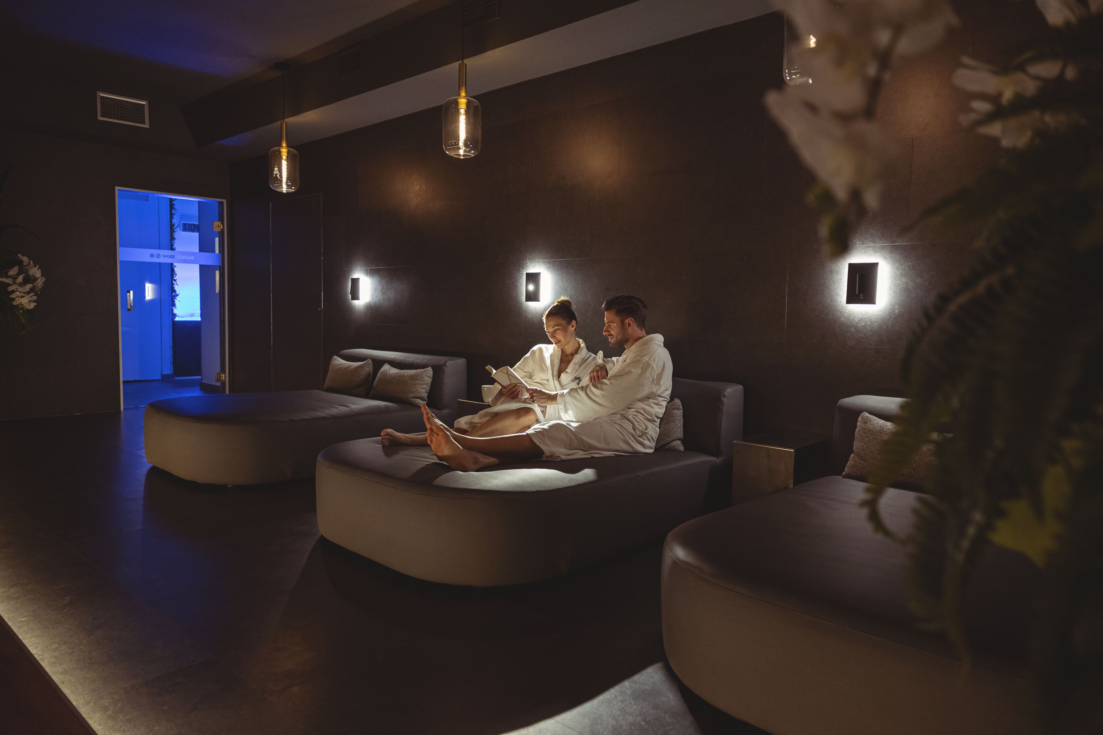 A modern spa area with a couple relaxing on a curved sofa, a reception desk, and a blue-lit entrance.