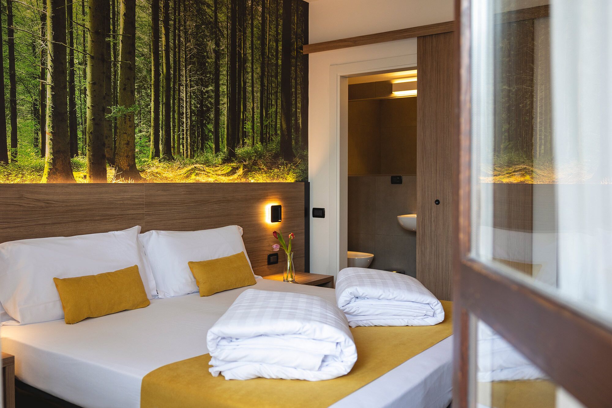 Ein Hotelzimmer mit einem Bett, einem Nachttisch, einer Vase mit Blumen und einem Wandbild eines Waldes.