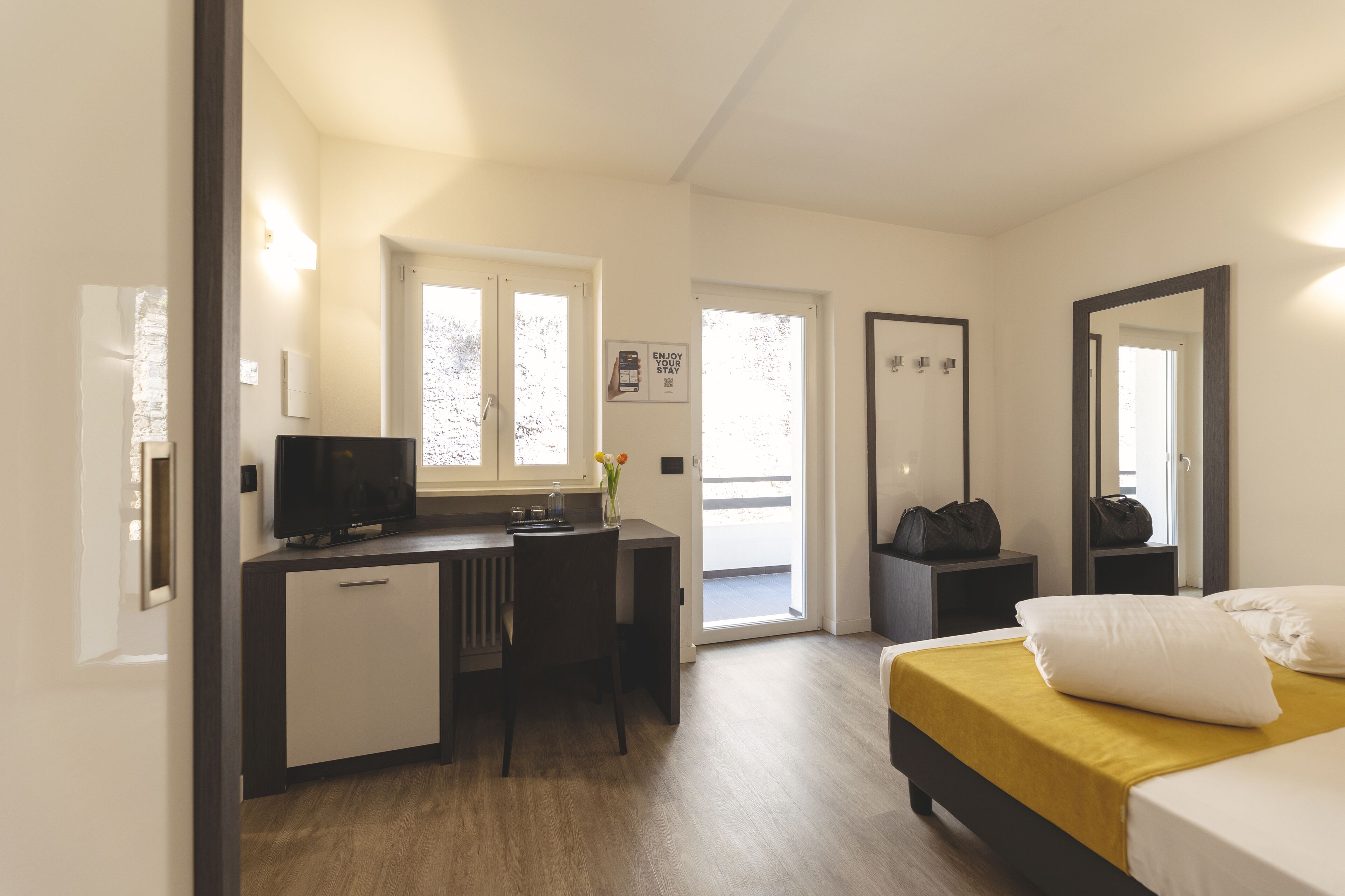 A modern hotel room with a bed, a desk with a computer, a chair, a small table, and a window.