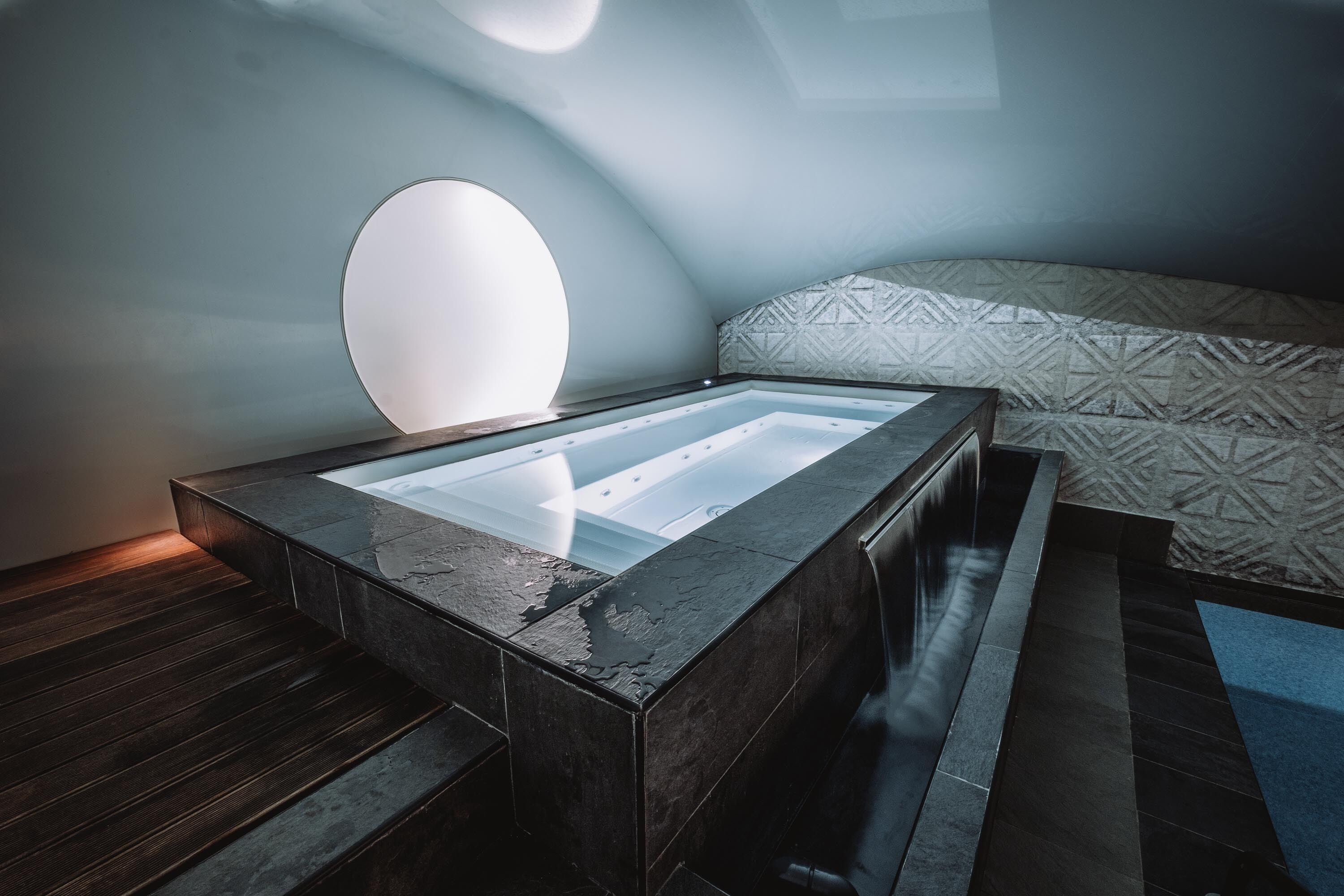 A modern bathroom with a glass-enclosed bathtub, a wooden floor, and a patterned wall.