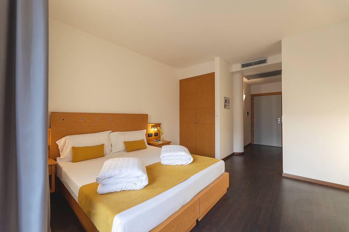 A hotel room with a large bed, a wardrobe, a bedside table with a lamp, and a door leading to another room.