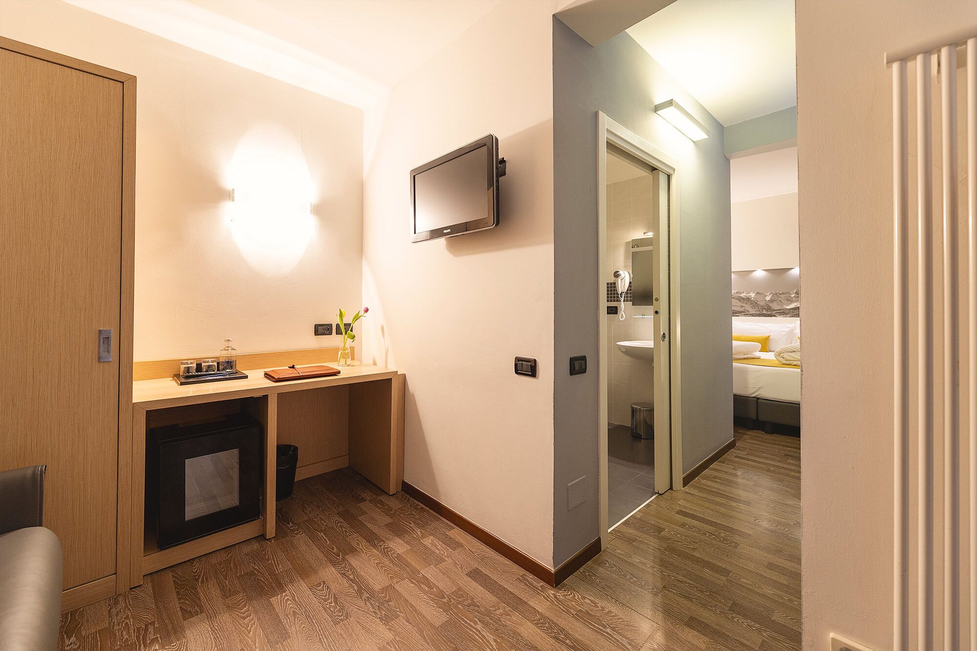 A hotel room with a wooden desk, a mirror, a television, and a bed with a mountain view.