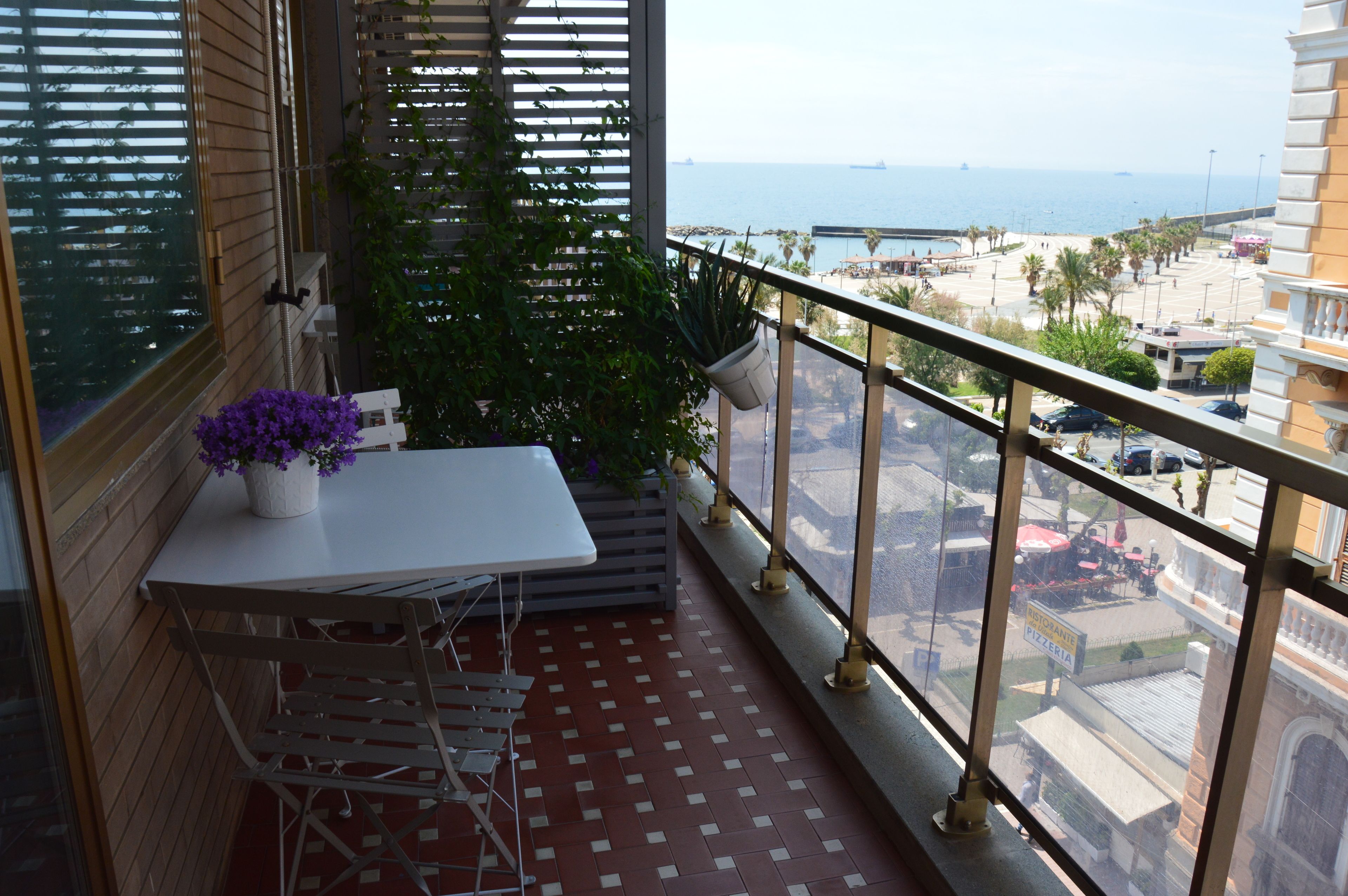 A balcony with a table and chairs, potted plants, and a view of the sea and cityscape.