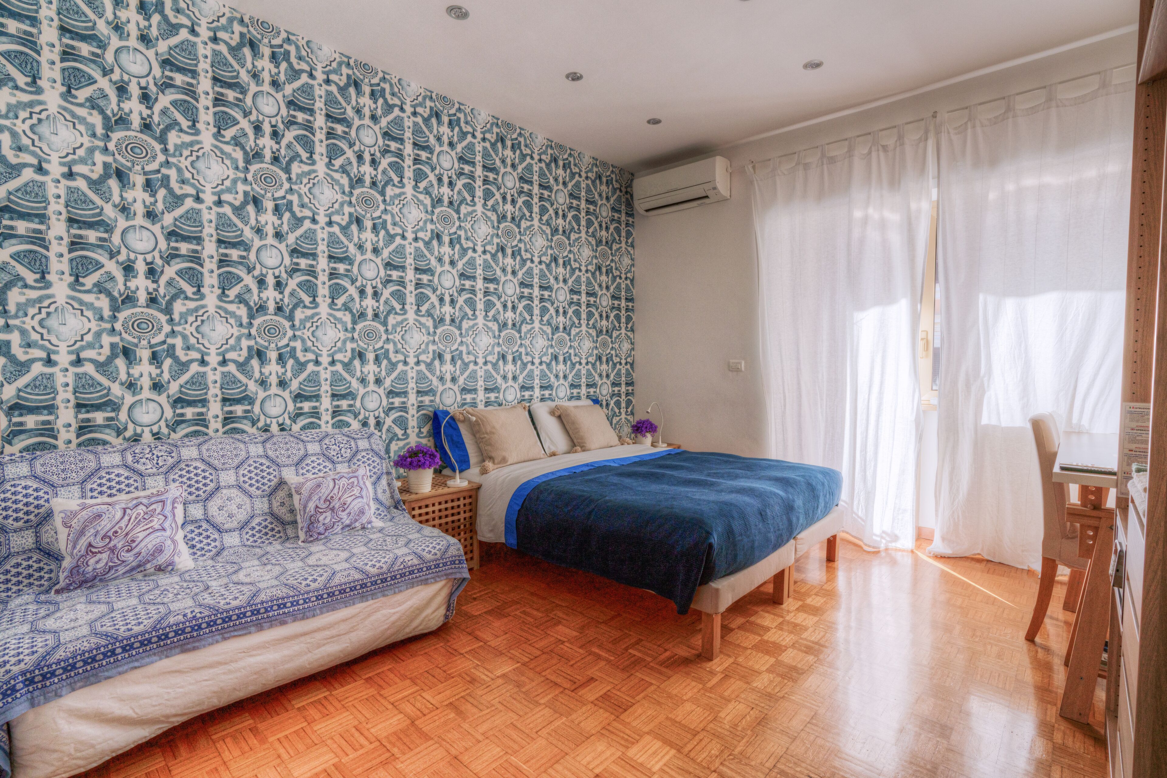 A bedroom with a patterned wall, a bed, a sofa, and a desk.