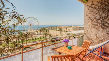 A balcony with a wooden table and chairs, a potted plant, and a view of a beach and a Ferris wheel.