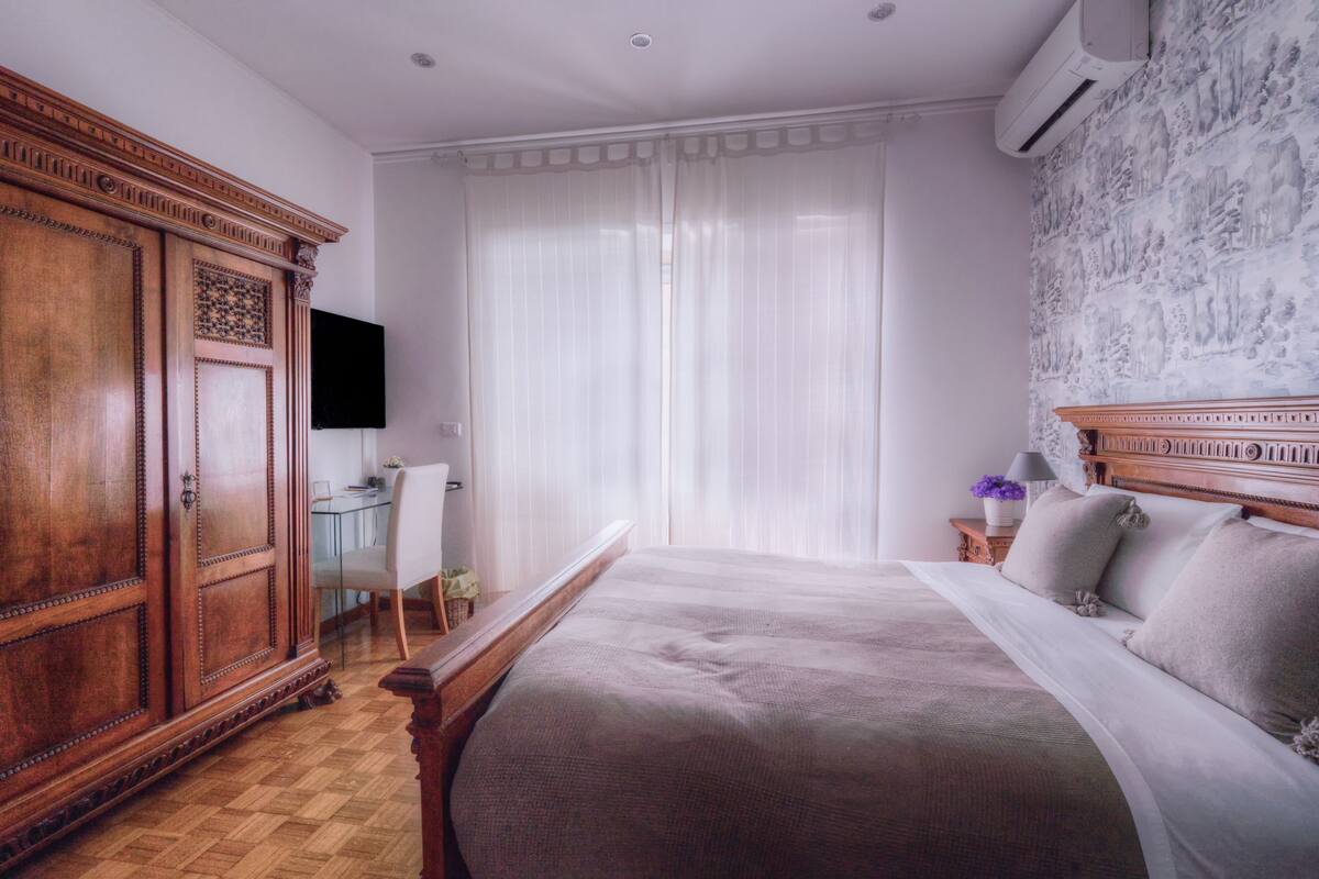 A bedroom with a large bed, a wooden wardrobe, a desk with a chair, and a wall-mounted TV.