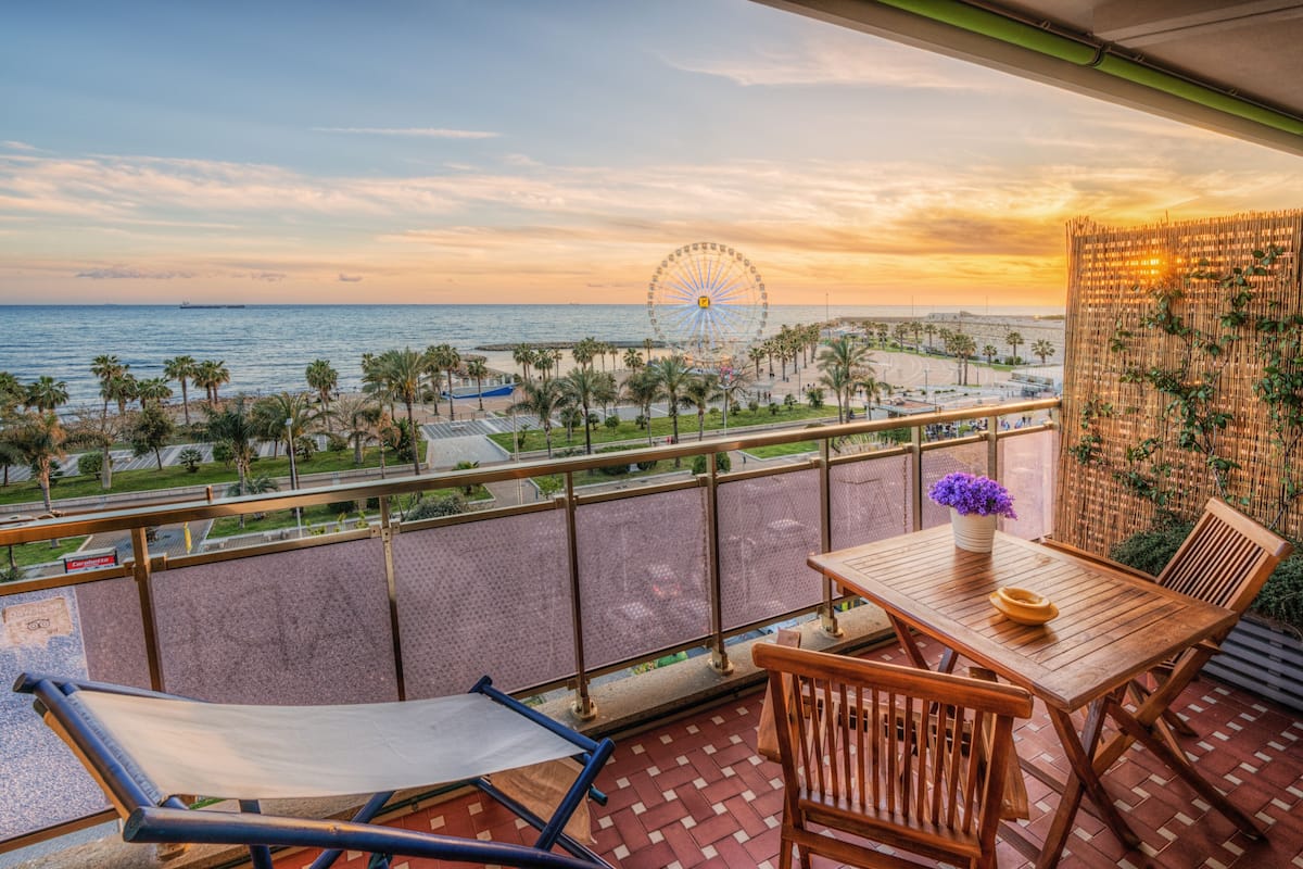 A balcony with a view of the sea and a Ferris wheel, wooden furniture, and a decorative wall.
