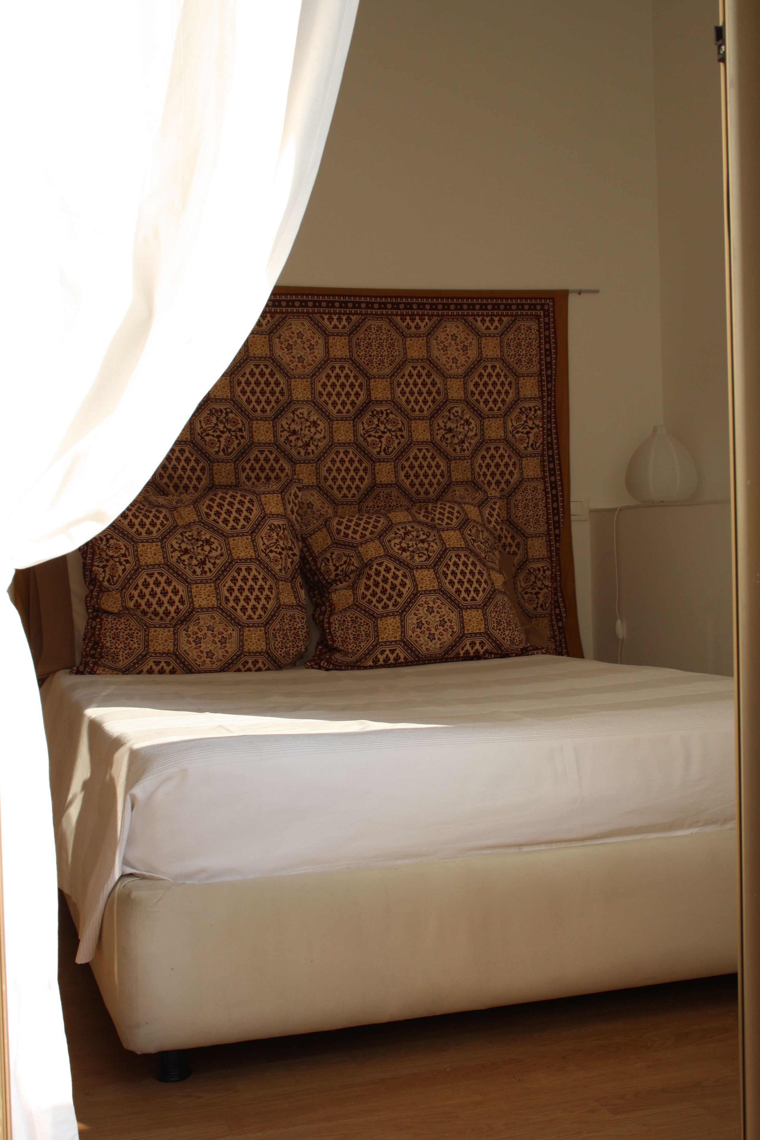 A bed with a patterned headboard, a bedside lamp, and a curtain partially drawn.