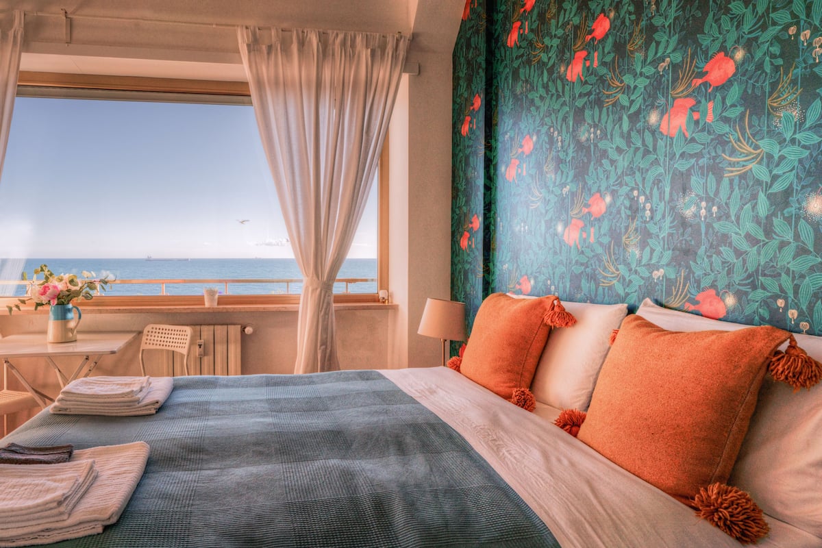 A hotel room with a bed, a view of the sea, a balcony with a table and chairs, and a window with curtains.