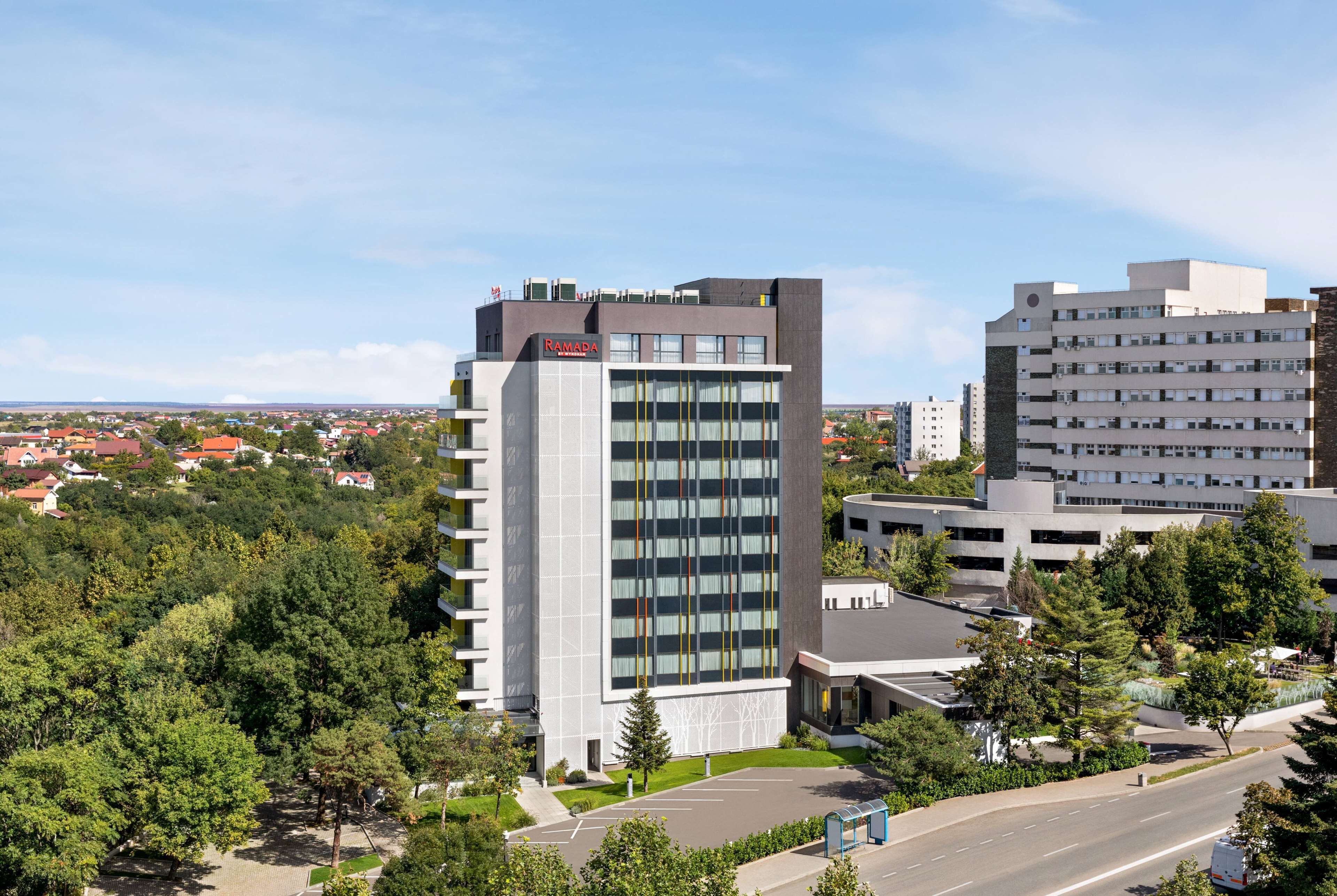 Moderni rakennus, jonka yläpuolella on suuri, huomiota herättävä «RAMADA»-kyltti. Rakennuksen ympärillä on vehreää kasvillisuutta ja taivaalla ei ole pilviä.