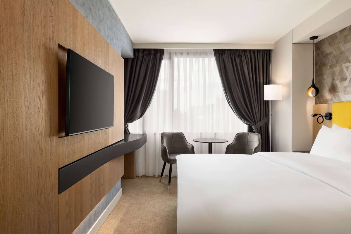 Una habitación de hotel moderna con una cama grande, un televisor de pantalla plana en la pared, una pequeña mesa redonda con dos sillas y un amplio ventanal con cortinas.