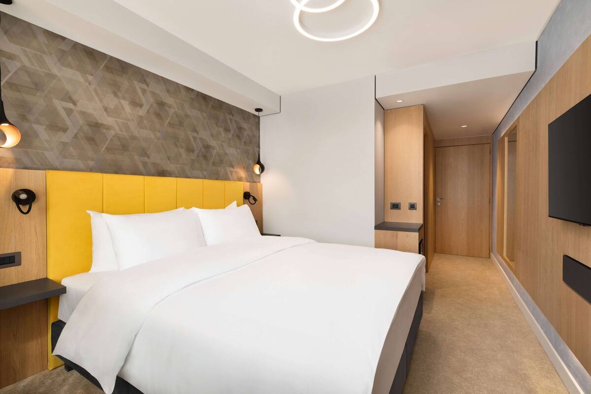 Una habitación de hotel moderna con una cama grande, un cabecero de madera, un televisor de pantalla plana en la pared y un banco incorporado al pie de la cama.