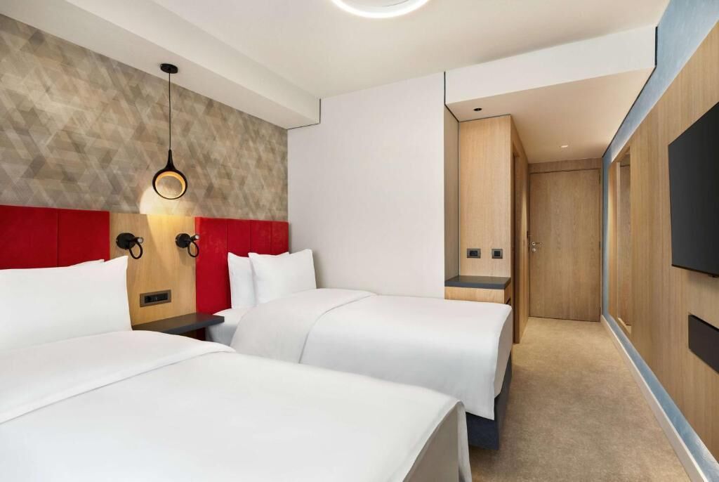 A hotel room with two beds, a red accent wall, a wooden headboard, a wall-mounted TV, and a modern light fixture.