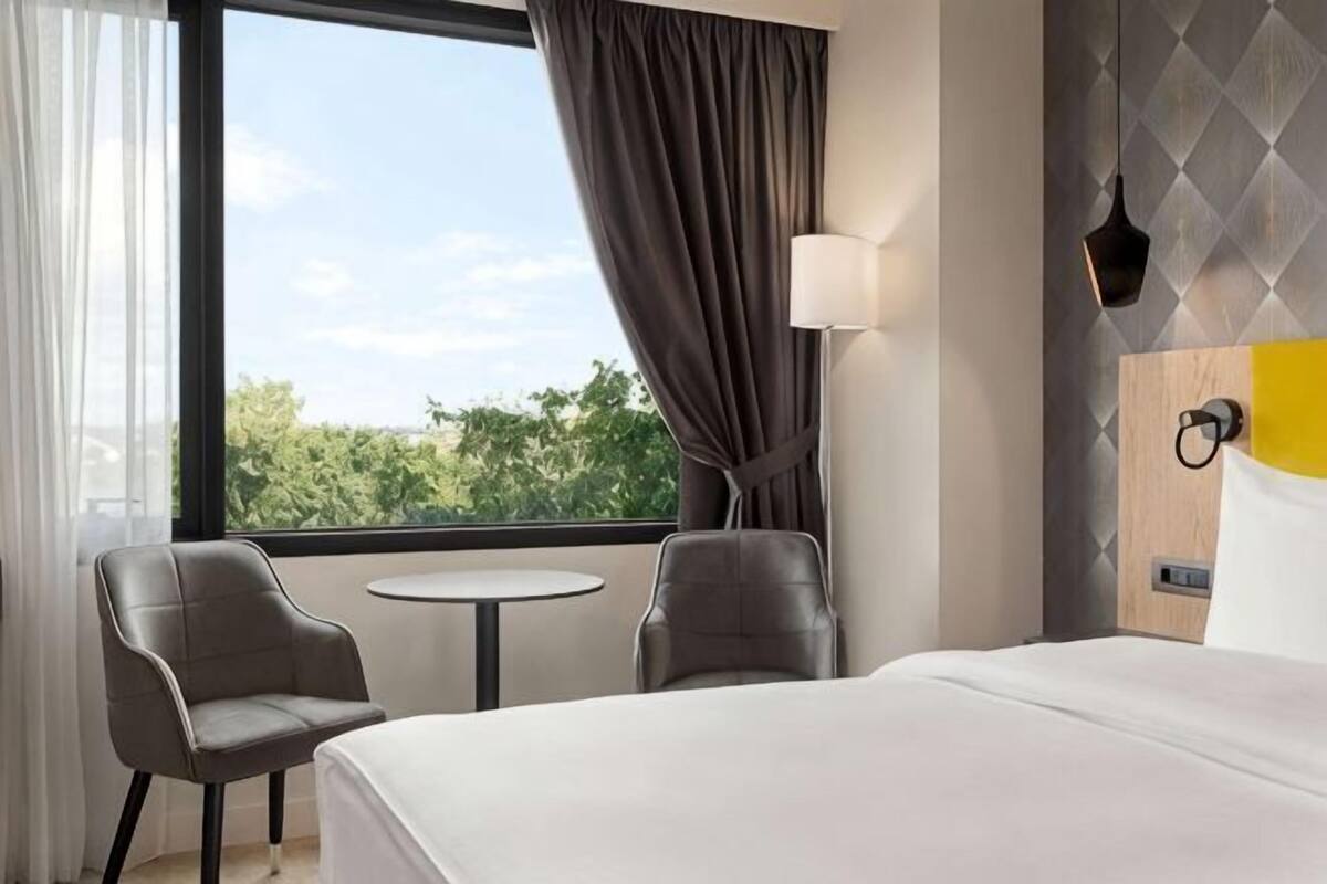 Habitación de hotel con cama, dos sillas, una mesita, ventana con cortinas y una lámpara de pared.