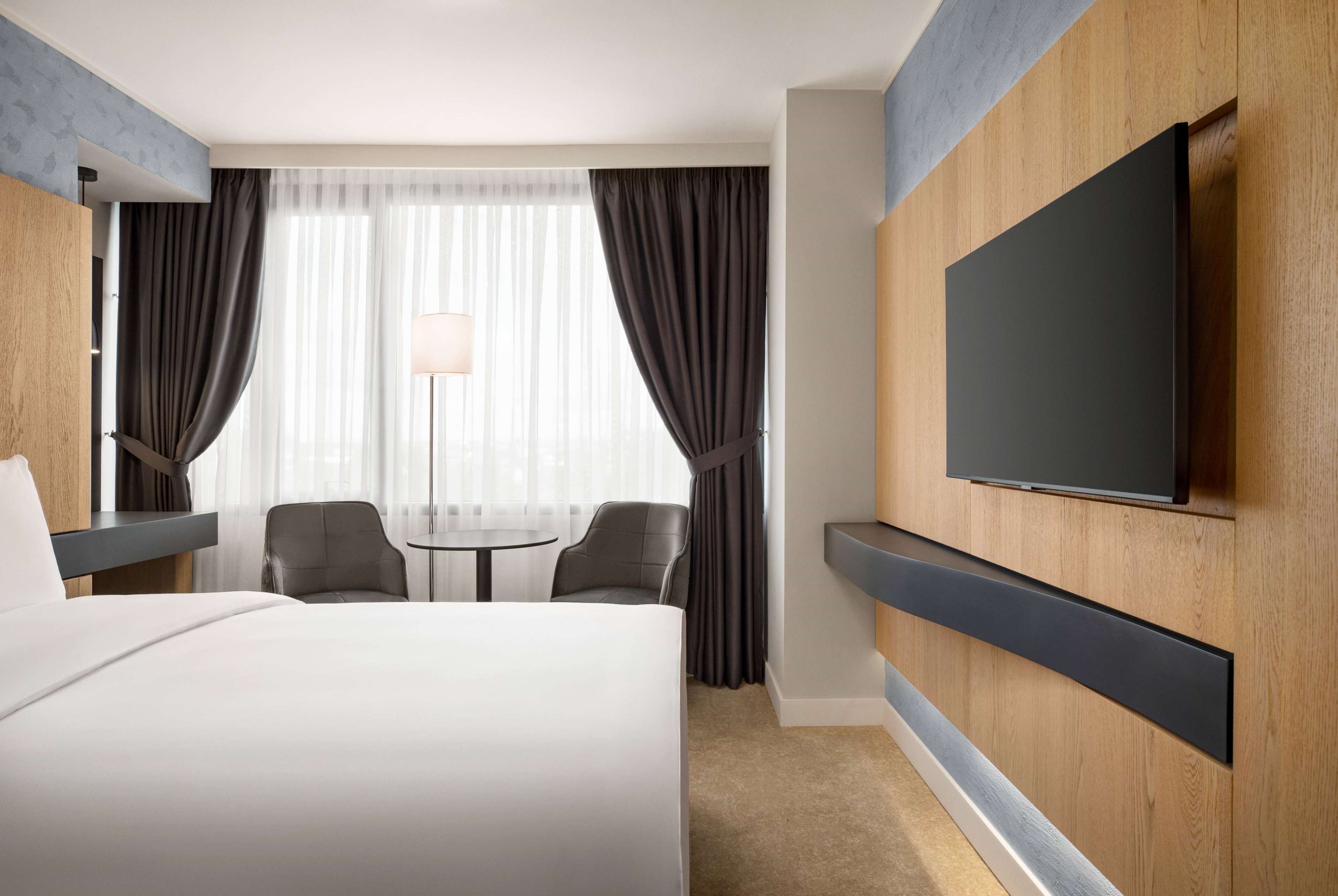Una habitación de hotel con una cama grande, un televisor montado en la pared, un escritorio con una silla y una ventana con cortinas.