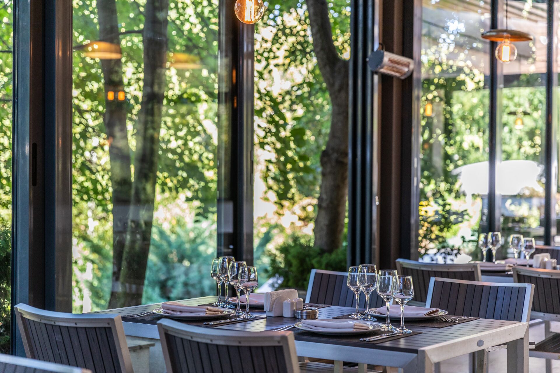 Un restaurant avec des tables dressées pour le service, de grandes fenêtres et des luminaires suspendus.