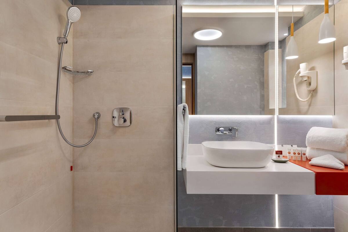 Un baño moderno con ducha a ras de suelo, un espejo grande y un lavabo blanco con encimera roja.
