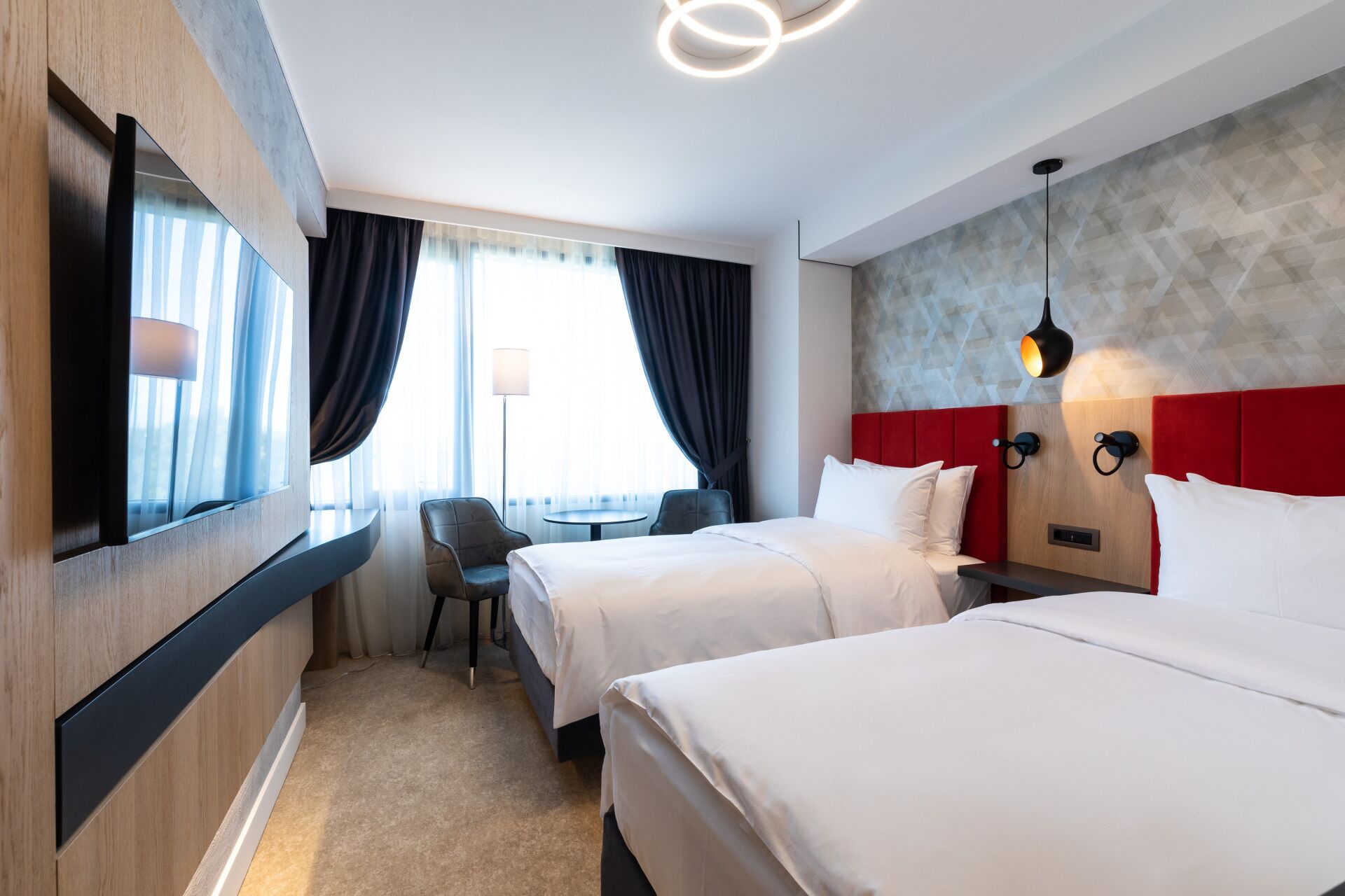 A hotel room with two beds, a desk, a chair, and a large window with curtains.