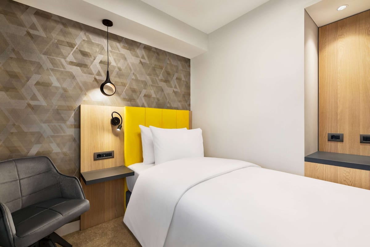 Una habitación de hotel moderna con una pared de diseño geométrico, un cabecero amarillo, una cama blanca, un sofá negro y una mesita de noche de madera con una lámpara.