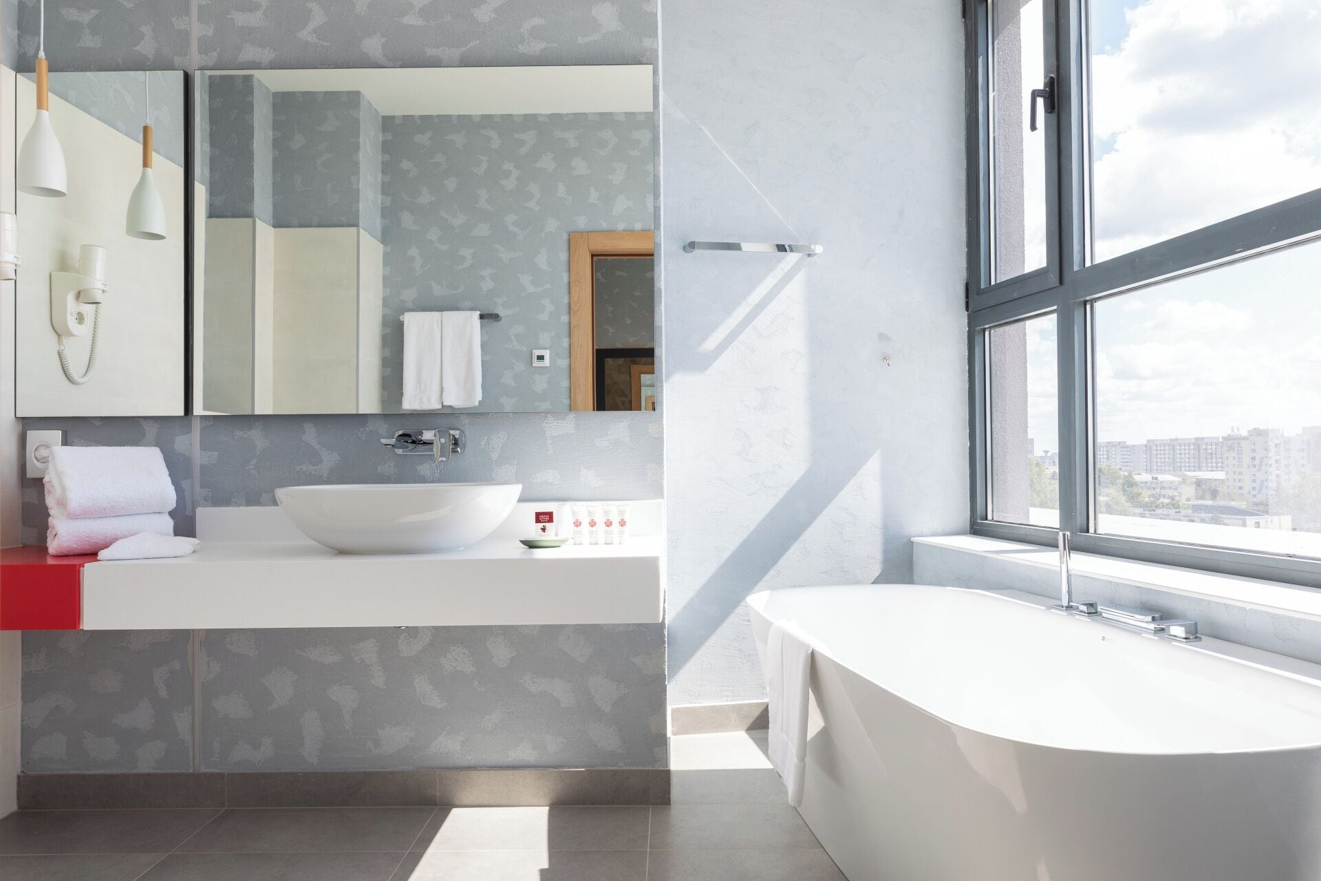 A modern bathroom with a large window, a freestanding bathtub, and a vanity with a large mirror.