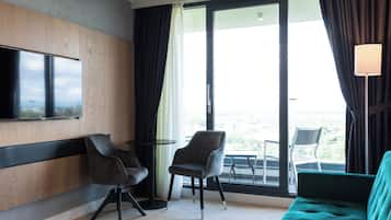 A hotel room with a large window, a flat-screen TV mounted on the wall, two chairs, a small round table, and a teal sofa.