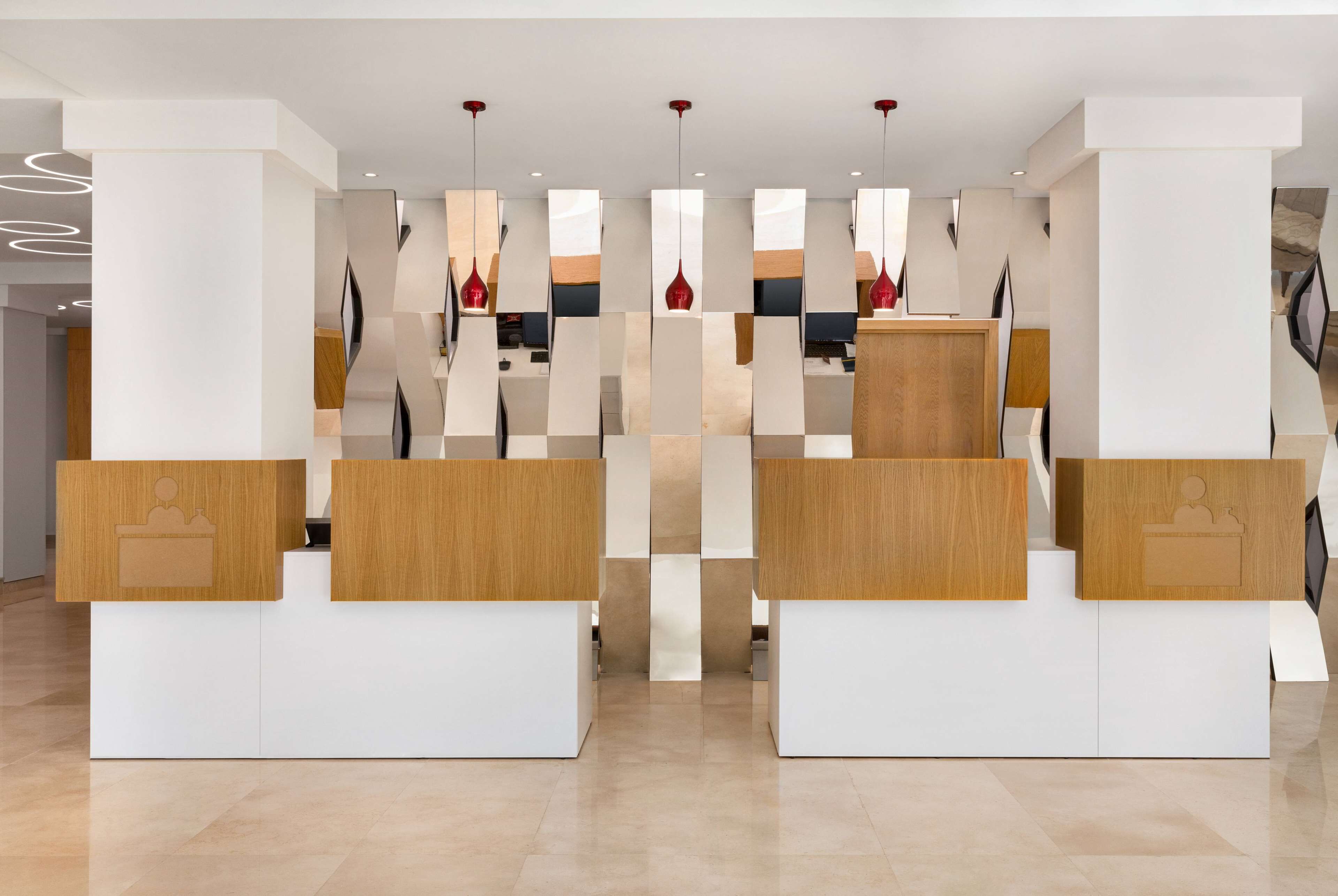 A reception area with modern design, featuring a central desk with a wooden finish and a white base. There are multiple red pendant lights hanging from the ceiling, and the floor is made of light-colored tiles.