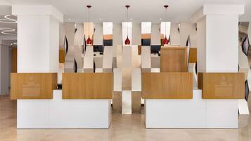 A reception area with modern design, featuring a central desk with a wooden finish and a white base. There are multiple red pendant lights hanging from the ceiling, and the floor is made of light-colored tiles.