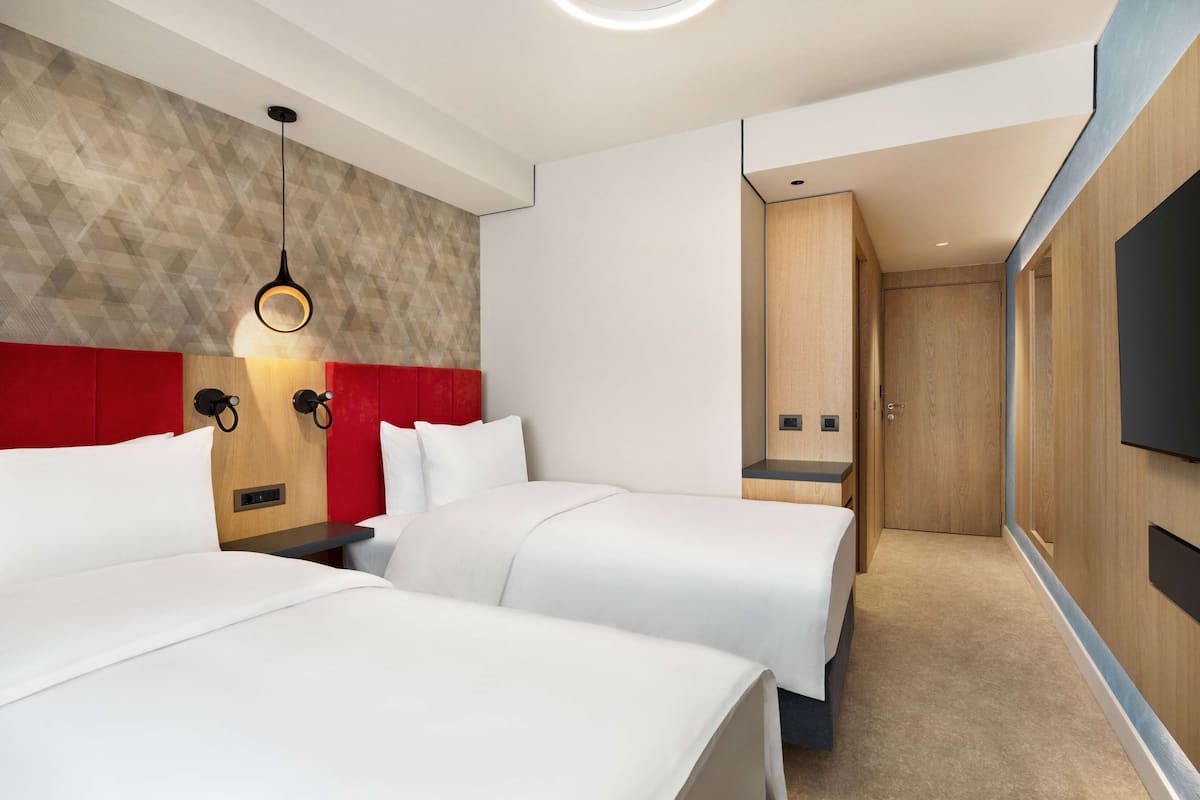 Habitación de hotel con dos camas, una pared de acento roja, un cabecero de madera, un televisor montado en la pared y una puerta de madera.