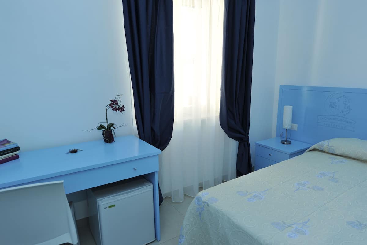 Ein Schlafzimmer mit Bett, Schreibtisch, kleinem Kühlschrank, Nachttisch und einem Fenster mit Vorhängen.
