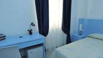 Un dormitorio con cama, escritorio, refrigerador pequeño, mesita de noche y ventana con cortinas.