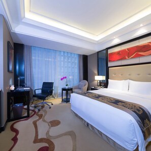 Une chambre d’hôtel avec un grand lit, un bureau avec une chaise, une petite table avec une lampe et une grande fenêtre avec des rideaux.