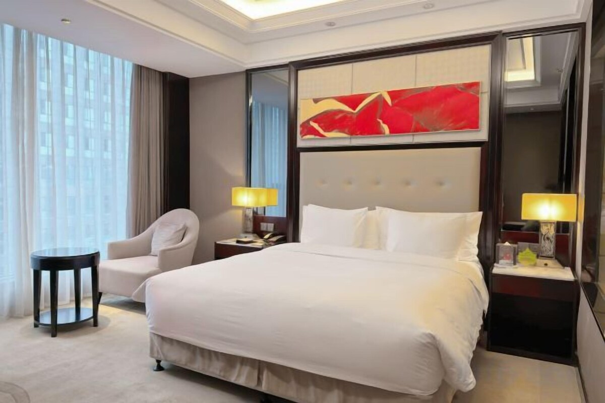 A hotel room with a large bed, two bedside tables with lamps, a chair, and a large window with curtains.