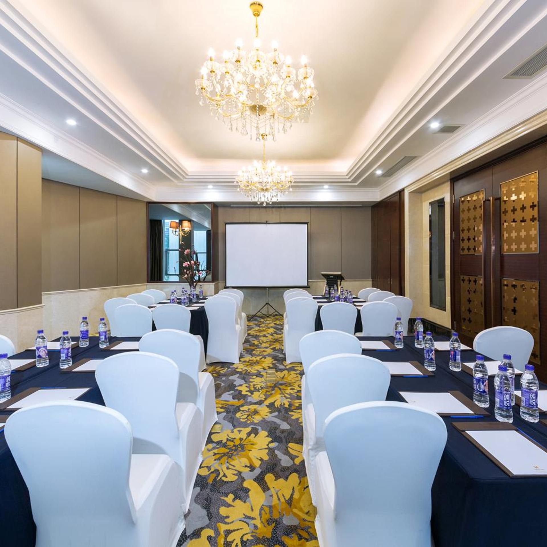 A conference room with a large screen, white chairs, water bottles, and a chandelier.