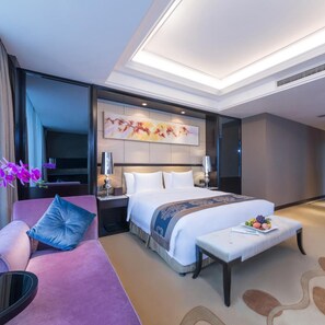 A hotel room with a large bed, a bedside table, a chair, a small table with fruit, and a decorative wall piece. - Dading Century Plaza Hotel (Chengdu)