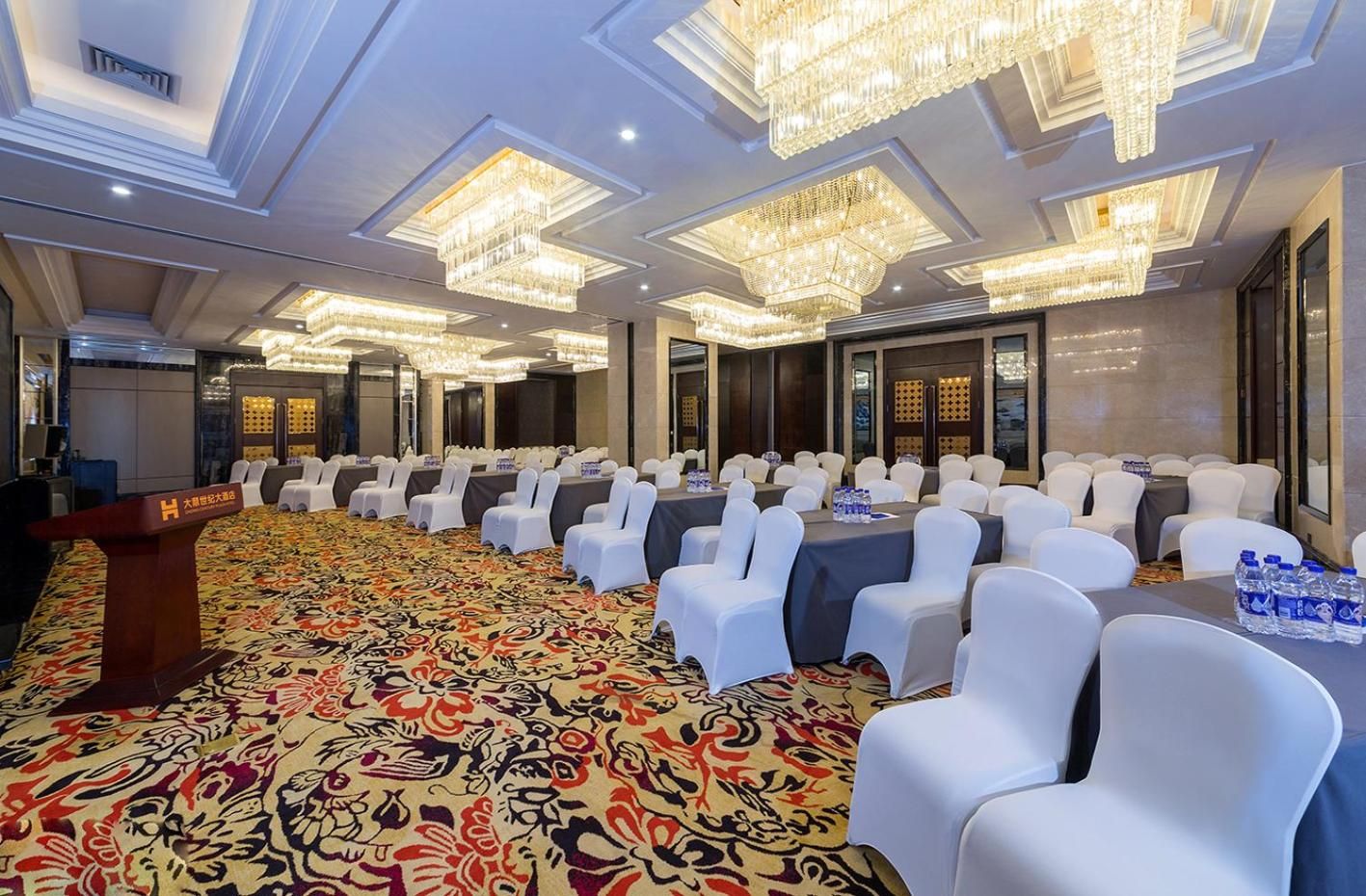 Sala de conferências com fileiras de cadeiras brancas, um púlpito e um tapete estampado.