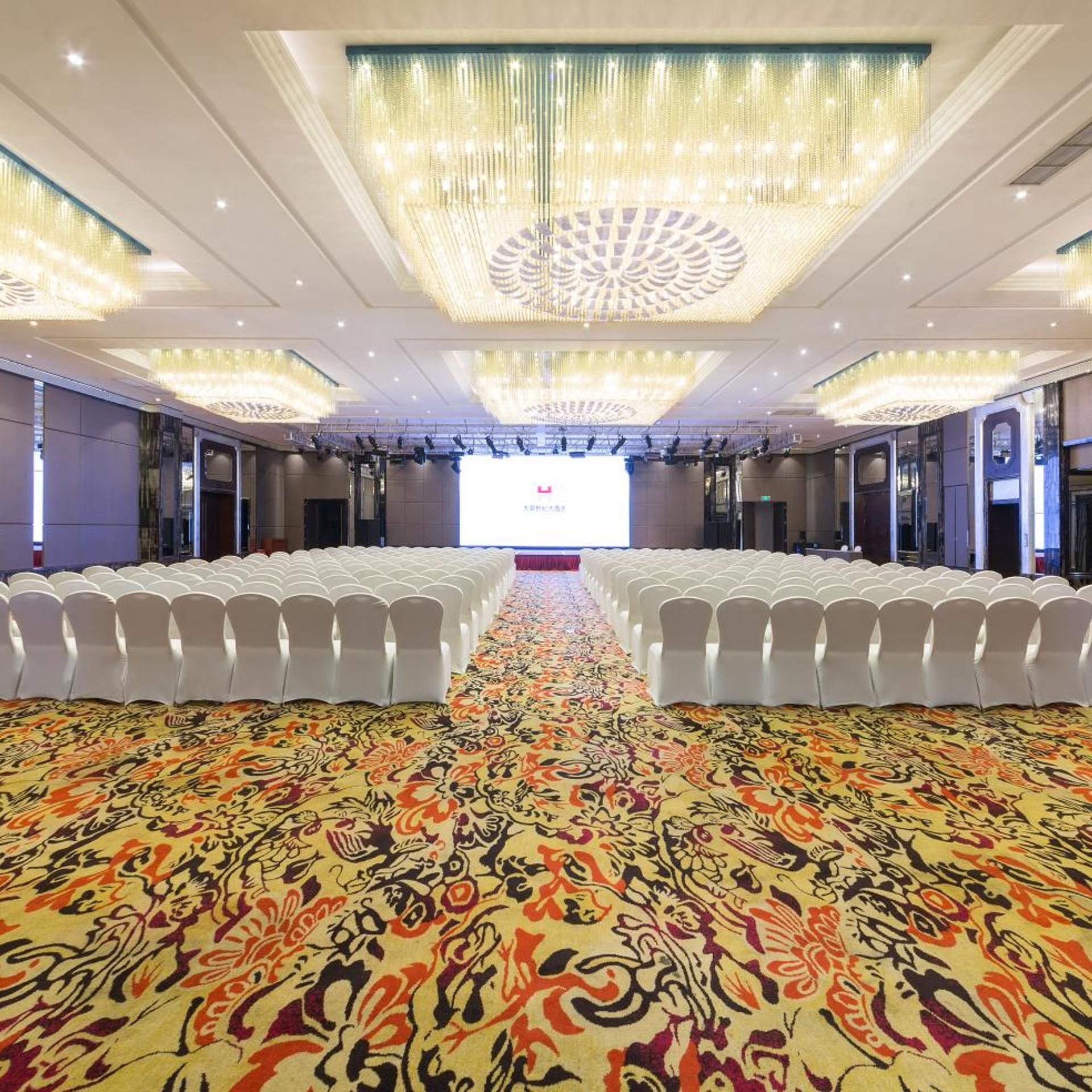 Un salón de conferencias con filas de sillas blancas, una alfombra estampada y una pantalla grande al frente.