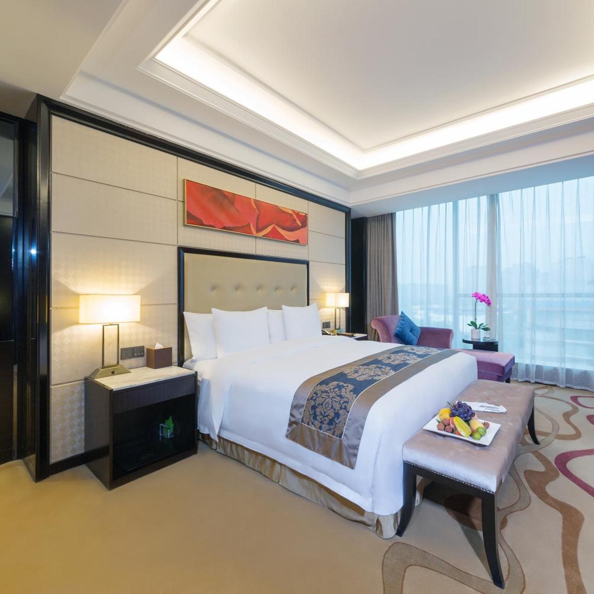 A hotel room with a large bed, bedside tables, a desk, and a view of the city through large windows.