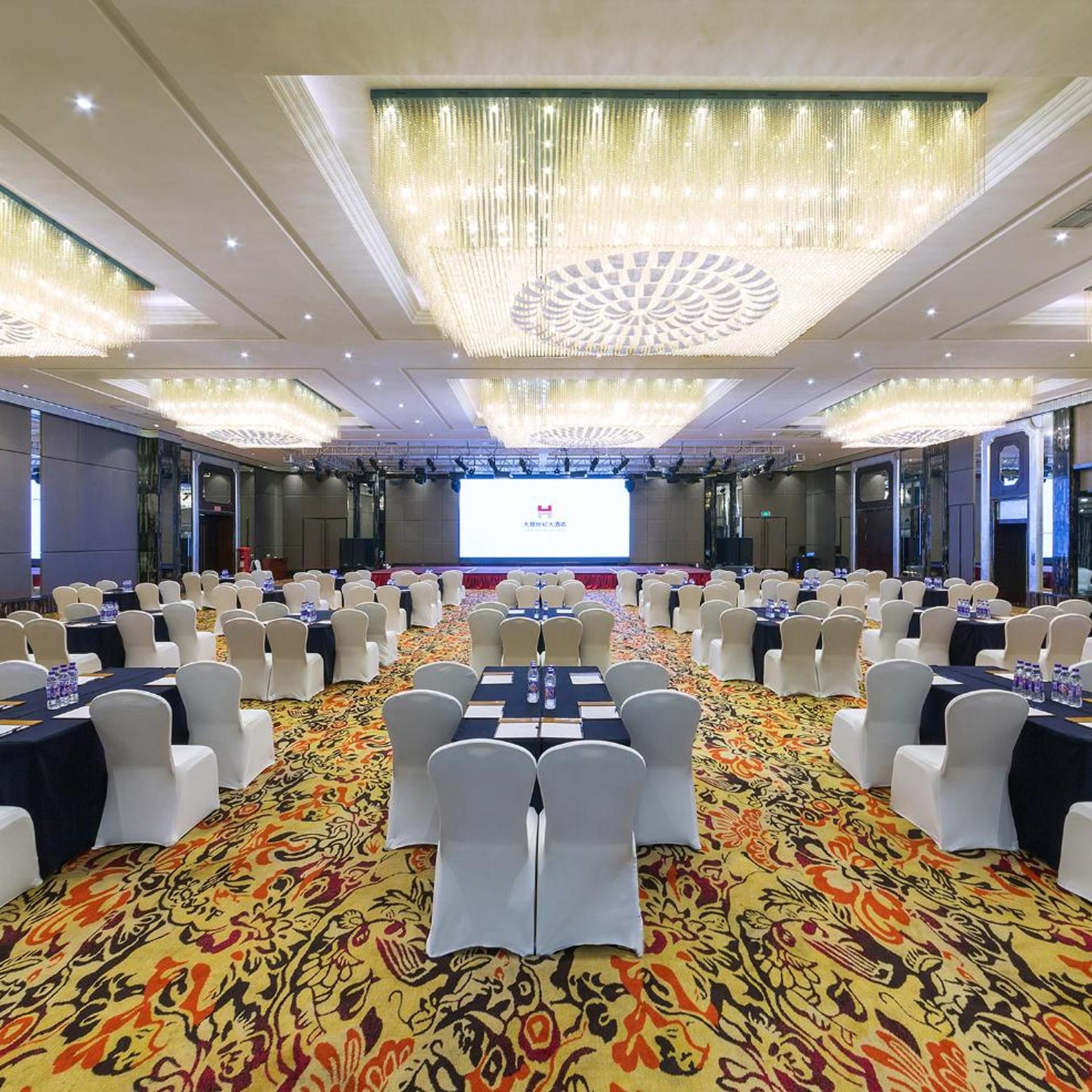 A conference room with rows of tables and chairs, a large screen at the front, and a patterned carpet.