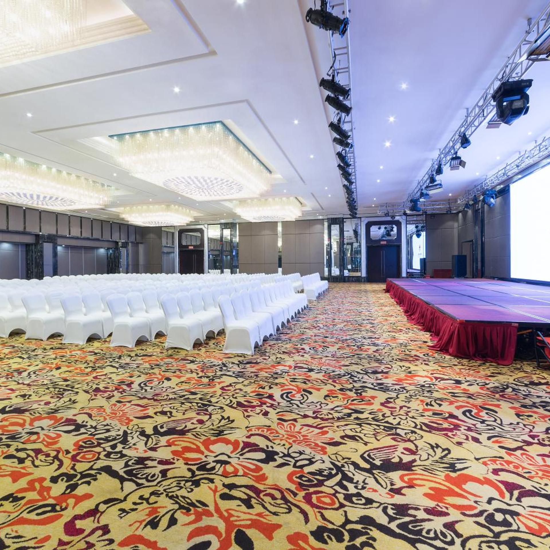 Un salón de conferencias con filas de sillas blancas, un escenario con un telón de fondo rojo y una alfombra estampada.