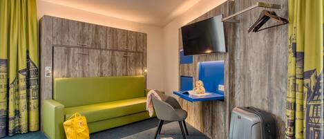 Habitación de hotel con un sofá verde, un escritorio pequeño con una televisión de pantalla plana, una maleta y un bolso amarillo.