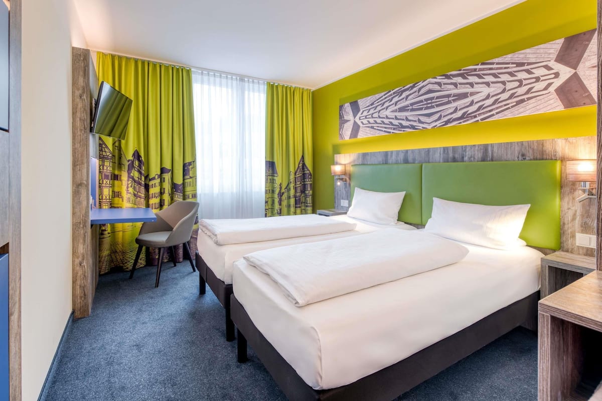 Ein Hotelzimmer mit zwei Betten, einem Schreibtisch, einem Stuhl und einem großen Fenster mit Vorhängen.