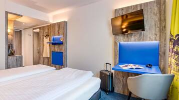 A hotel room with a bed, a desk with a chair, a TV mounted on the wall, and a suitcase on the floor.